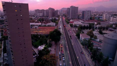 A-subway-train-moving-through-a-bustling-cityscape-at-dusk,-framed-by-buildings,-aerial-view