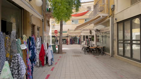 Una-Calle-Estrecha-En-Una-Ciudad-Chipriota-Llena-De-Tiendas-Que-Exhiben-Telas-Coloridas-En-El-Exterior,-Con-Algunos-árboles-Y-Asientos-Al-Aire-Libre.