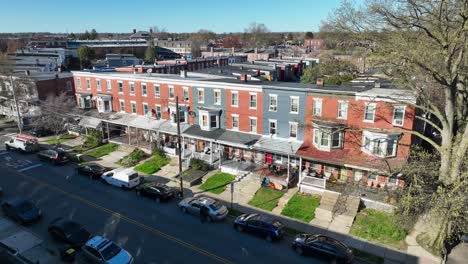 Aerial-view-of-a-residential-street,-row-of-red-brick-townhouses-with-white-trim-and-front-porches,-lined-with-parked-cars-on-both-sides-under-a-clear-sky
