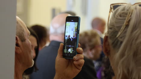 Person-recording-a-Spanish-communion-on-a-smartphone-in-a-crowded-church-hall,-vibrant-and-lively-atmosphere