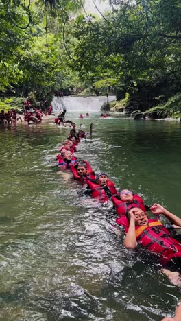 Menschen-Tragen-Sicherheitswesten-Und-Schweben-Im-Urlaub-In-Einer-Reihe-Auf-Dem-Gebirgsfluss-Mit-Wasserfall-Im-Hintergrund