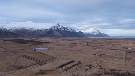 Felder-Und-Landwirtschaftliche-Gebäude-In-Island-Landschaft-Unter-Schneebedeckten-Bergen