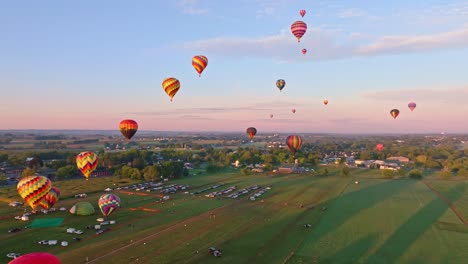 Eine-Flotte-Lebhafter-Heißluftballons-Erhebt-Sich-Sanft-In-Einen-Warmen-Sonnenuntergangshimmel-über-Einer-Ruhigen-Ländlichen-Landschaft