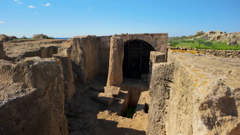 A-gated-entrance-leads-to-ancient-burial-chambers-at-the-Tombs-of-the-Kings,-set-against-a-backdrop-of-extensive-stone-ruins