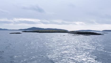 Passing-view-from-passenger-ferry-of-rugged,-rocky-and-small-islands-with-birds-and-South-Uist-in-the-distance-in-the-Outer-Hebrides-of-Scotland-UK