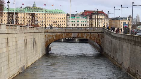 Puente-Sobre-El-Canal-De-Agua-Con-Arquitectura-De-Gran-Hotel-En-El-Fondo-En-Estocolmo,-Suecia,-Cámara-Lenta