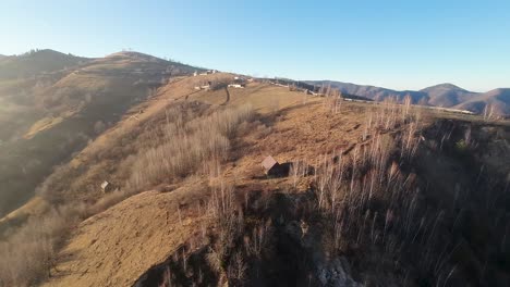 Aerial-view-of-a-mountainous-landscape-in-late-autumn,-showcasing-scattered-cabins,-barren-trees,-and-rolling-hills-under-a-clear-sky