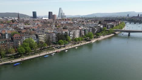 An-aerial-perspective-of-Basel,-Switzerland's-residential-area-nestles-gracefully-alongside-the-flowing-Rhine-River,-symbolizing-the-harmonious-coexistence-between-urban-living-and-natural-waterways