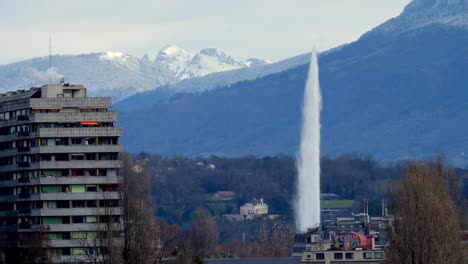 Una-Toma-Del-Jet-D&#39;eau-En-Ginebra,-Con-Un-Edificio-A-La-Izquierda-De-La-Toma-Y-Los-Alpes-Al-Fondo