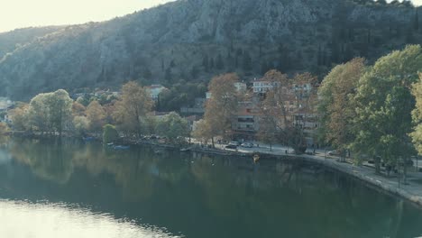 Drone-FLIGHT-aside-to-a-village-on-a-hill-in-Europe,-green-trees,-sunny-weather,-blue-water-lake,-SCENIC-mountains,-Greece-4K-video