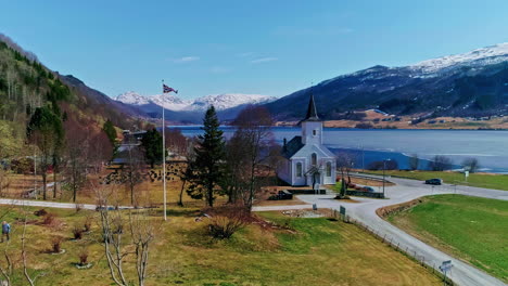 Norwegian-Countryside-Scenery,-Aerial-View-of-Church,-Coastal-Road,-Ice-on-Fjord-Water-on-Sunny-Day