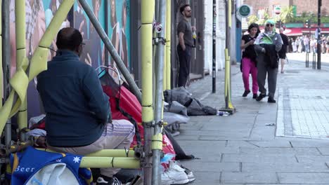 A-homeless-man-sits-smoking-a-cigarette-in-between-scaffolding-where-a-second-person-lies-in-a-red-tent-surrounded-by-people’s-belongings-as-members-off-the-public-pass-by