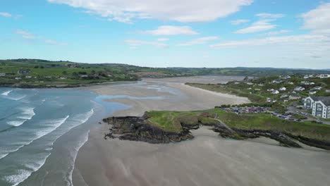 Aerial-footage-over-sandy-beach-Inchidoney-on-low-tide-with-waves-and-green-hills-panorama,-village-with-houses-and-rugged-coastline