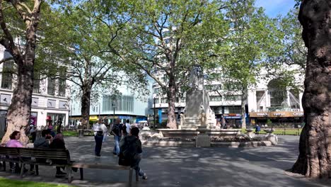 Leicester-Square-On-Sunny-Morning-With-People-Relaxing-On-Benches-And-Takings-Photos-Of-Statue-of-William-Shakespeare