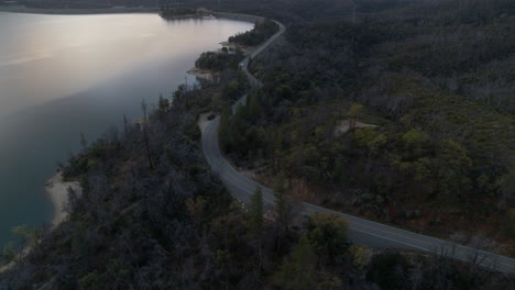 Panorámica-Aérea-Sobre-Una-Carretera-Que-Sigue-Los-Contornos-Del-Lago-Whiskytown-En-El-Norte-De-California-Con-Un-Camión-Circulando-Por-Ella.