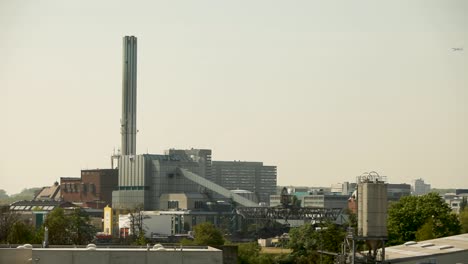 Urban-industrial-landscape-with-tall-chimney-and-airplane-flying-in-the-background,-warm-daylight