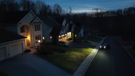 Twilight-aerial-shot-of-a-suburban-street,-featuring-well-lit,-spacious-houses-with-peaked-roofs-and-garages,-and-a-car-parked-in-street