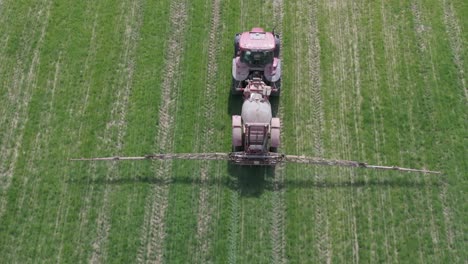 Vuelo-De-Drones-Detrás-De-Un-Tractor-Rojo-Rociando-Con-Su-Remolque-En-Un-Campo-De-Cultivos-Alimentarios,-Nos-Acercamos-Gradualmente-Al-Tractor-Tomando-Un-Primer-Plano