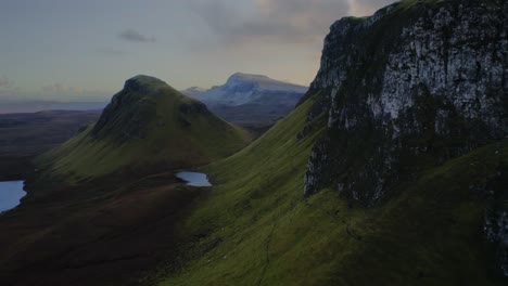 Flight-near-rocky-cliffs-unveils-lush-green-highlands,-lakes,-and-a-distant-mountain-at-Quiraing-Walk,-Isle-of-Skye,-Scotland