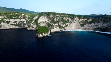 Drone-flying-over-blue-sea-toward-Diamond-Beach,-Nusa-Penida,-Bali-in-Indonesia
