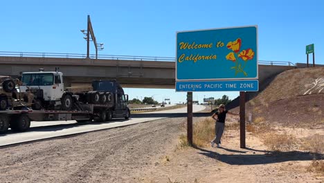 Young-blonde-woman-posing-for-picture-under-Welcome-to-California-state-sign