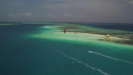 A-red-kite-surfing-near-cayo-sardina,-vibrant-turquoise-waters-and-boats,-aerial-view