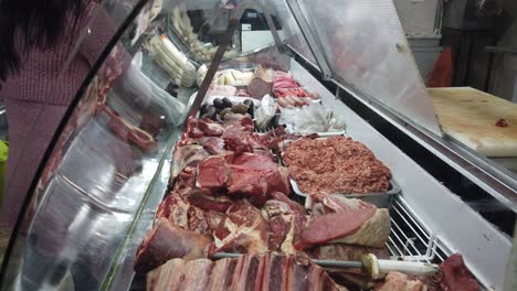 Carnicero-Seleccionando-Cortes-De-Carne-En-Una-Concurrida-Tienda-De-Asados-Argentinos,-Mostrando-Variedad-En-Una-Exhibición-Bien-Iluminada.