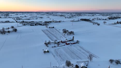 Vista-Aérea-De-Una-Granja-Cubierta-De-Nieve-Al-Atardecer,-Que-Muestra-Graneros-Y-Silos-Rojos,-Rodeados-De-Vastos-Campos-Abiertos-Y-Nevados-Bajo-Un-Cielo-Pastel.