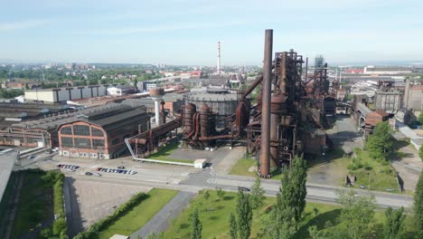 Vista-Aérea-De-Una-Antigua-Planta-Industrial-Con-Grandes-Estructuras-Metálicas-Oxidadas,-Rodeada-De-Vegetación-Y-Una-Carretera,-Bajo-Un-Cielo-Despejado