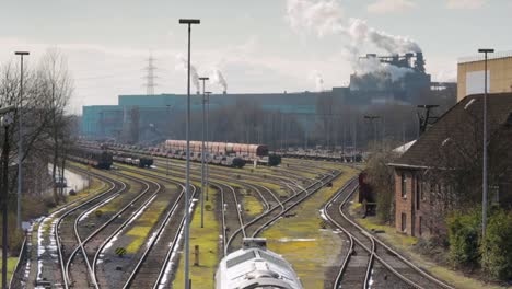 Concurrido-Patio-Ferroviario-Con-Múltiples-Trenes-De-Carga,-Edificios-Industriales-Al-Fondo-Emitiendo-Humo,-Día-Soleado