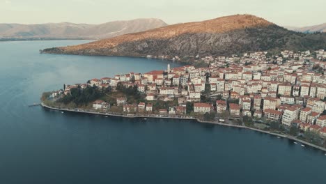 Drone-EPIC-FLIGHT-above-a-village-on-a-hill-in-Europe,-red-roof-tops,-little-houses,-sunrise-weather,-blue-water-lake,-SCENIC-mountains,-Greece-4K-video