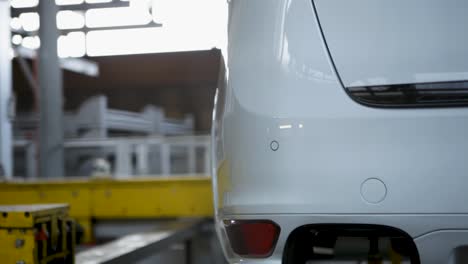 Coche-Blanco-En-Un-Ascensor-En-Un-Taller-De-Automoción,-Fondo-Ligeramente-Borroso-Con-Un-Técnico-Trabajando