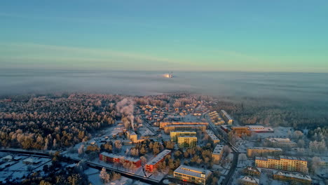 Vista-Aérea-De-Una-Pequeña-Ciudad-Industrial-En-Un-Soleado-Día-De-Invierno,-Edificios-Nevados-Y-Bosques-Con-Una-Central-Eléctrica-De-Carbón-En-Un-Horizonte-Brumoso