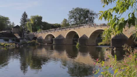 Sunlit-ancient-stone-bridge-over-calm-river-with-reflections-and-greenery,-clear-blue-sky