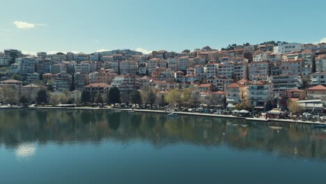 Drones-Volando-A-Un-Lado-De-Un-Pueblo-En-Una-Colina-Verde-En-Europa,-Tejados-Rojos,-Clima-Soleado,-Lago-De-Agua-Azul,-Video-De-4k-En-Grecia