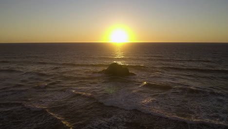 Sun-sets-on-horizon-directly-above-single-rock-in-ocean-battered-by-waves-as-spray-rises,-Oregon-Coast