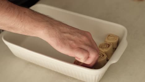 White-man-placing-homemade-cinammon-buns-into-white-ceramic-baking-tray-on-kitchen-countertop