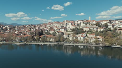 Drone-flying-towards-a-village-on-a-green-hill-in-Europe,-red-roof-tops,-little-houses,-sunny-weather,-blue-water-lake,-Greece-4K-video,-zoom-in-movement