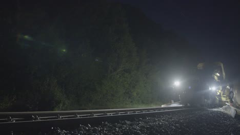 Toma-Nocturna-De-Maquinaria-De-Mantenimiento-Ferroviario-En-Funcionamiento,-Iluminada-Por-Luces-Brillantes,-Rodeada-De-Un-Denso-Follaje