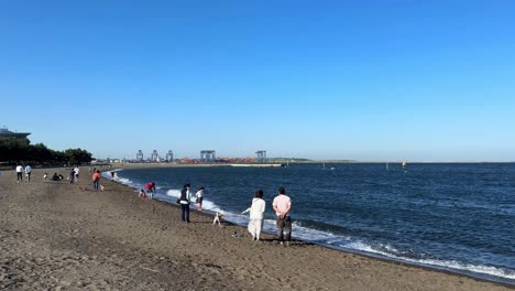 Beach-scene-with-people-strolling-and-fishing-on-a-sunny-day,-industrial-port-in-background,-calm-ocean-waves