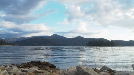 Static-camera-on-the-rocky-beach-of-a-lake-with-morning-clouds-slowly-moving-over-the-mountains-in-the-background