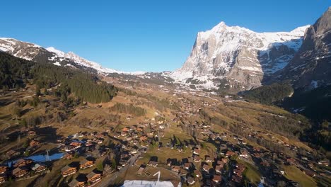 Impresionante-Vista-Aérea-Del-Pueblo-Alpino-En-Primavera-Con-Picos-Montañosos-Cubiertos-De-Nieve-En-El-Fondo