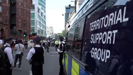 A-group-of-Metropolitan-police-officers,-including-a-chief-inspector,-stand-next-to-a-dark-blue-Territorial-Support-Group-riot-van-as-other-officers-clear-the-road-during-a-public-order-event