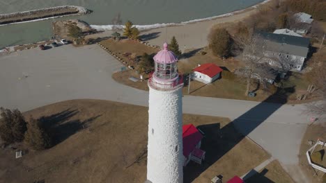 A-scenic-lighthouse-with-pink-top-on-a-sunny-day,-surrounded-by-trees-and-near-a-body-of-water,-aerial-view