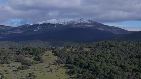 Vuelo-Lateral-Con-Dron-Creando-Un-Efecto-De-Paralaje-Aparece-Nieve-La-Montaña-Con-El-Nombre-Pico-De-El-Mirlo-Se-Mueve-Por-Una-Ladera-Verde-Hay-Nubes-En-La-Cumbre-Gredos-Valle-Del-Tietar-España