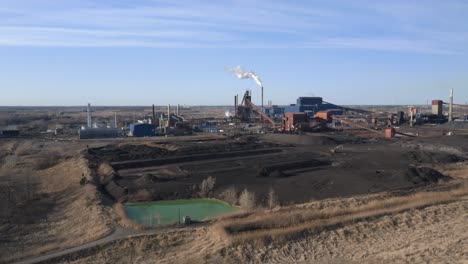 Wide-angle-view-of-a-large-industrial-site-with-multiple-structures,-smokestacks-emitting-steam,-and-a-clear-sky