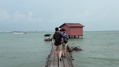 Dolly-in-of-people-walking-on-the-wooden-bridge-and-against-the-beautiful-background-view-of-the-iconic-Red-House-at-Tan-Jetty,-one-of-the-Clan-Jetties-at-Weld-Quay,-Georgetown-Penang,-Malaysia