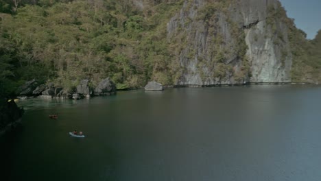 Tourists-in-canoes-in-calm-water-by-cliffs-in-Phillipinnes,-low-fast-forward-aerial