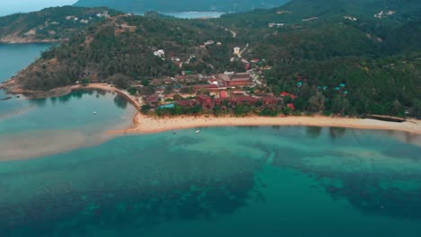 Aerial-drone-shot-of-a-tropical-beach-resort-surrounded-by-lush-greenery-and-crystal-clear-waters,-showcasing-a-serene-coastal-landscape-in-Thailand