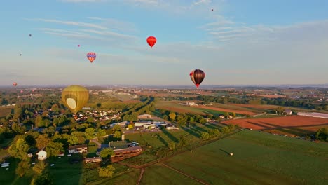 Vista-Aérea-De-Coloridos-Globos-Aerostáticos-Que-Se-Elevan-Hacia-El-Cielo-De-La-Mañana-Sobre-Un-Tranquilo-Paisaje-Rural